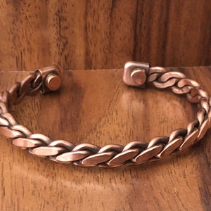 May include: A copper braided bracelet with magnetic clasps. The bracelet is made of a thick, braided copper wire and has a simple, elegant design.