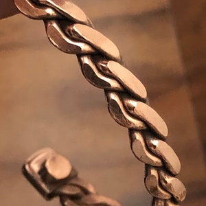 May include: A copper bracelet with a braided design. The bracelet is made of a single piece of copper that is twisted and woven together to create a unique and stylish look.