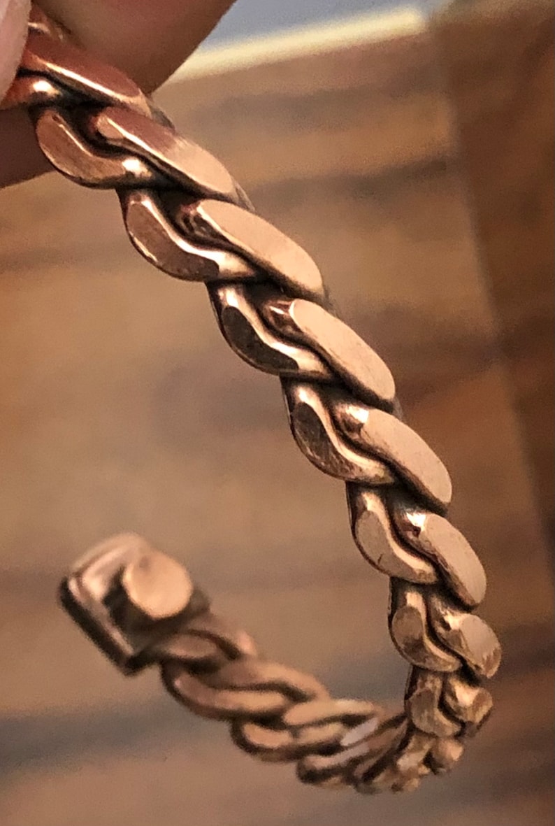 May include: A copper bracelet with a braided design. The bracelet is made of a single piece of copper that is twisted and woven together to create a unique and stylish look.