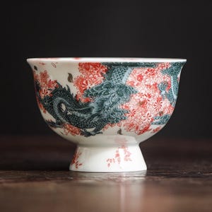 Op de afbeelding: Een witte keramische kom met een voetstuk, versierd met een gedetailleerd drakenontwerp in teal en rode bloempatronen. Het ontwerp van de kom wikkelt zich om de buitenkant, waardoor een visueel rijke esthetiek ontstaat. De kom wordt waarschijnlijk gebruikt om te serveren of tentoon te stellen.