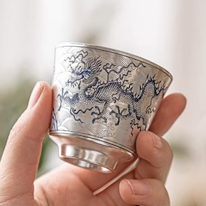 Könnte beinhalten: Eine silberfarbene Teetasse mit einem detaillierten blauen Drachendesign. Die Tasse hat einen ausgestellten Rand und einen kleinen, kreisförmigen Boden. Der Drache ist auf der Außenseite der Tasse aufwendig geätzt und erzeugt eine strukturierte Oberfläche.