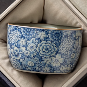 May include: A blue and white floral patterned ceramic bowl. The bowl is presented in a gift box with a soft, beige interior. The floral design features various flower types in shades of blue and white, creating a detailed and elegant aesthetic.