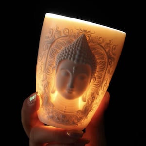 May include: A white ceramic candle holder with a detailed relief of Buddha's face and ornate patterns. The candle holder is illuminated from within, creating a warm, glowing effect against a dark background.