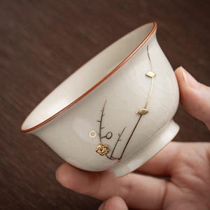 May include: A white ceramic teacup with a brown rim and a decorative floral design. The cup has a small footed base and is held in a hand. The floral design is in gold and black.