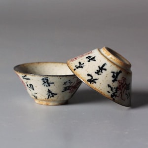 May include: Two small, off-white ceramic tea bowls with black and red calligraphy. One bowl is upright, the other is tilted, revealing a brown rim. The bowls have a speckled texture and a rustic aesthetic.