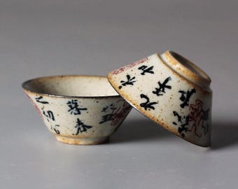 May include: Two small, off-white ceramic tea bowls with black and red calligraphy. One bowl is upright, the other is tilted, revealing a brown rim. The bowls have a speckled texture and a rustic aesthetic.