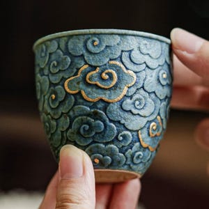 Puede incluir: Una pequeña taza de té de cerámica texturizada en un tono azul verdoso profundo, con un patrón en relieve de nubes estilizadas en su exterior. Algunos contornos de las nubes están resaltados con pintura dorada metálica, creando un contraste con la superficie mate.