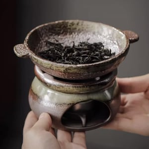 May include: A handcrafted ceramic tea warmer with a bowl-shaped top filled with loose tea leaves. The warmer has a multi-toned glaze in shades of brown, green, and gray. The tea warmer is held in a person's hands.
