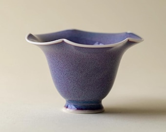 May include: A handcrafted, purple ceramic bowl with a unique, scalloped rim and a speckled texture. The bowl has a flared shape and a small, pedestal base. The interior is a darker shade of purple, and the rim is trimmed with white.