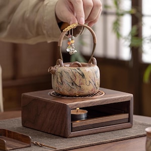 May include: A ceramic teapot with a decorative handle and a small charm is held above a wooden tea warmer. The teapot has a mottled beige and brown pattern. The warmer has a drawer for a small candle.