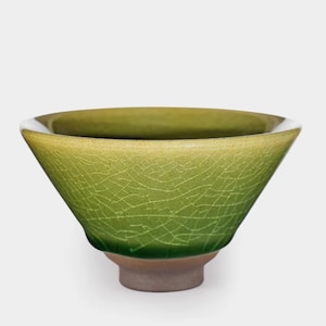 Può includere: Una ciotola in ceramica verde con smalto craquelé. La ciotola ha una forma ampia e angolata con un bordo bianco e una piccola base rotonda marrone. L'interno della ciotola è verde più scuro.