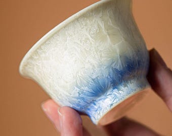 May include: A small, ceramic teacup with a unique crystalline glaze. The cup transitions from a light cream color at the top to a vibrant blue at the base. The glaze creates a textured, almost frosted appearance, adding visual interest to the piece.