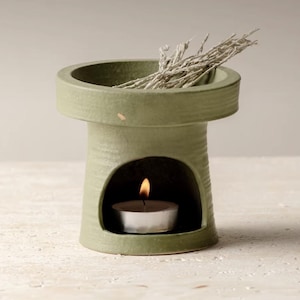 Puede incluir: Un quemador de aceite de cerámica verde salvia con una base cilíndrica y una parte superior ancha. Una pequeña vela de té arde dentro del calentador, y tallos secos se colocan en la parte superior. El calentador tiene un diseño minimalista.