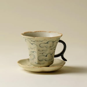 May include: A ceramic teacup and saucer set. The cup has a unique, fluted shape with a black handle and is decorated with a blue cloud pattern. The saucer is round and matches the cup's design. The set is off-white.