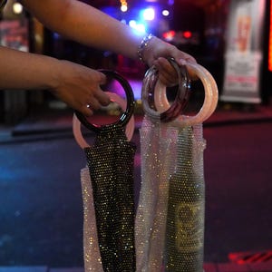 Puede incluir: Dos bolsas de malla con cuentas y asas circulares se sostienen en alto. Una bolsa es negra, la otra plateada. Una botella está dentro de una bolsa de malla plateada. Las asas son transparentes, negras y rosas.