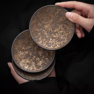 May include: A stack of three round, ceramic coasters with a mottled, metallic bronze and gold finish. The coasters are held by a person's hand against a black background. The top coaster is being lifted, revealing the stack below.