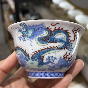Pode incluir: Chávena de chá de cerâmica branca com um desenho de dragão azul. O dragão é representado com escamas azuis e detalhes vermelhos, num fundo de ondas azuis. A base da chávena apresenta um padrão geométrico.