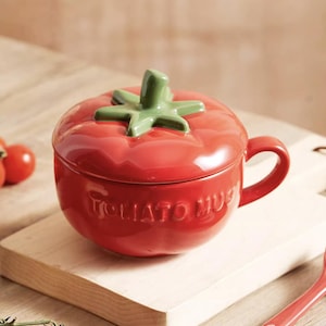 May include: A red ceramic tomato-shaped mug with a green stem-like handle and lid. The mug has the words "TOMATO MUG" embossed on the side. The mug is sitting on a wooden cutting board with a few tomatoes.