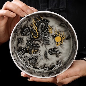 May include: A white ceramic bowl featuring a detailed black and gold dragon design. The dragon is depicted amidst swirling clouds and waves, with a golden sun. The bowl is held by hands.