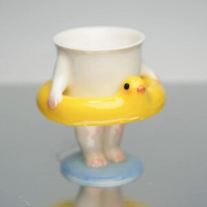 Könnte beinhalten: Eine skurrile Keramiktasse mit weißem Körper, rosa Armen und Füßen sowie einem gelben, entenförmigen Schwimmring. Die Tasse steht auf einem blau-weißen Sockel und schafft ein verspieltes Design. Ein lustiges und dekoratives Objekt.