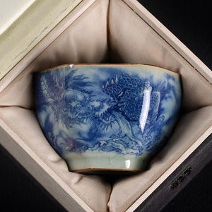 Può includere: Una tazza da tè in ceramica blu e bianca con un motivo a drago, presentata in una scatola foderata di beige. La tazza da tè presenta una dettagliata illustrazione di un drago in tonalità blu su sfondo bianco. La tazza ha una forma unica e una base verde chiaro.