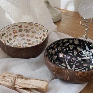 Puede incluir: Dos cuencos decorativos de coco con interiores de mosaico. Un cuenco presenta un mosaico de piezas de tonos neutros, mientras que el otro tiene un interior negro con acentos iridiscentes. Un manojo de palitos de madera de color claro descansa en primer plano.