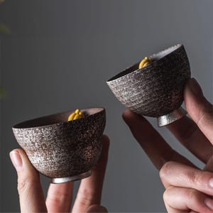 May include: Two small, handcrafted ceramic bowls with a textured, speckled brown exterior and a silver base. The bowls have a unique, asymmetrical shape and contain a small amount of yellow material. The bowls are held in front of a neutral background.