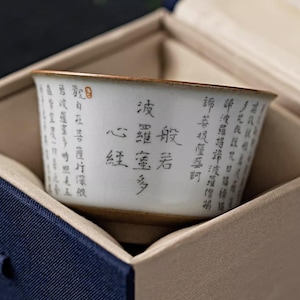 May include: A white ceramic teacup with black calligraphy and a brown rim, presented in a blue and beige box. The cup features intricate Chinese characters, suggesting a focus on traditional tea culture and aesthetics.