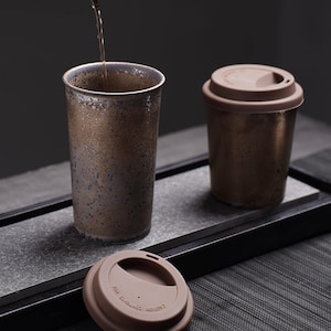 Może przedstawiać: Zestaw trzech brązowych ceramicznych kubków podróżnych z pokrywkami. Jeden wysoki kubek jest napełniany ciemnym płynem. Pozostałe dwa kubki są krótsze, jeden z pokrywką, a drugi bez. Kubki mają teksturowane, nakrapiane wykończenie.