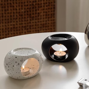 May include: Two ceramic essential oil diffusers. One is white with black speckles, the other is black. Both have a circular opening with a lit tea light candle inside. The diffusers sit on a white surface.
