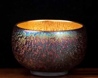 Könnte beinhalten: Eine kleine, schillernde Schale mit strukturierter Oberfläche. Die Außenseite der Schale zeigt ein Farbspektrum, darunter Lila, Grün und Gold. Das Innere ist goldfarben. Die Schale steht auf einem kleinen, grauen Sockel und ruht auf einer Holzoberfläche.