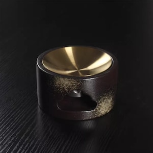 May include: A cylindrical, dark brown ceramic oil warmer with a gold-colored metal top. The warmer has a small opening on the side and a speckled, aged appearance. The background is a dark, solid color.