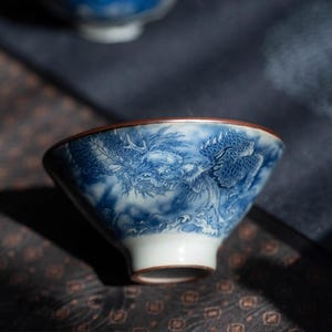 May include: A blue and white porcelain tea bowl with a dragon design. The bowl has a flared shape with a brown rim and a white base. The dragon is depicted in a detailed, traditional style, with clouds in the background.