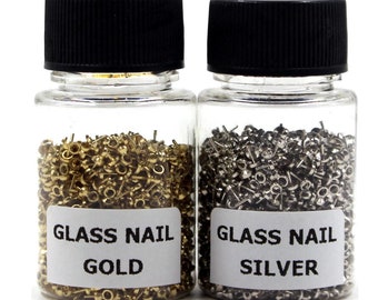 Könnte beinhalten: Zwei kleine, durchsichtige Glasflaschen gefüllt mit goldenen und silbernen Glasnägeln. Die Flaschen haben schwarze Deckel und weiße Etiketten mit der Aufschrift "Glass Nail Gold" und "Glass Nail Silver".