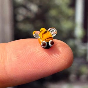 May include: A tiny glass bee figurine with yellow and black stripes and black eyes. The bee is sitting on a finger.