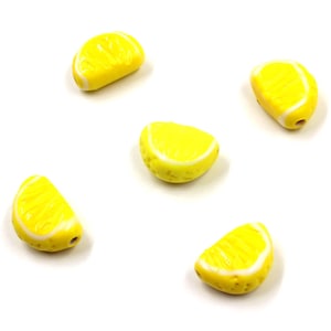 May include: Five yellow lemon slice beads with white rind details are scattered on a white background. These beads are designed to resemble a lemon slice. The beads have a hole for stringing.