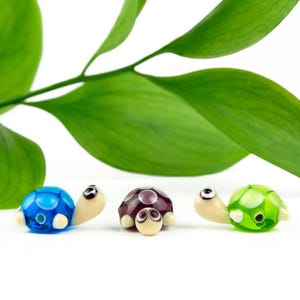 May include: Three small glass turtle figurines arranged in a row. The left turtle is bright blue, the center turtle is deep purple with white spots, and the right turtle is lime green. Each turtle has a cream-colored head with black eyes.