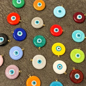 May include: A collection of twenty-one circular glass charms, each featuring a central eye motif with concentric rings of blue, white, and black. The charms come in a variety of solid colors including red, orange, yellow, green, blue, pink, and white.