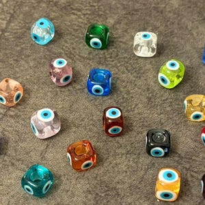 May include: A collection of seventeen small, square-shaped glass beads, each featuring a centered evil eye design. The beads are rendered in a variety of translucent and opaque colors, including blue, green, red, orange, yellow, purple, and clear.