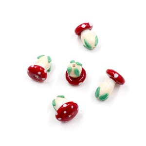 Op de afbeelding: Vijf rode en witte glazen kralen in de vorm van een paddenstoel met groene bladeren. De kralen zijn voor het maken van sieraden.