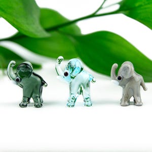 Op de afbeelding: Drie miniatuur glazen olifantenbeeldjes in groen, blauw en grijs. Elke olifant heeft een omhoog geheven slurf en kleine zwarte ogen. De achtergrond heeft groene bladeren.