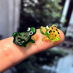 以下が含まれることがあります： 2つの小さなガラス製のカエルの置物。1つは濃い緑色、もう1つは黒い斑点のある黄色です。どちらも大きな白い目を持ち、指の上に置かれています。カエルは小さく、細部までこだわって作られており、手作りのようです。
