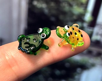 May include: Two miniature glass frog figurines. One is dark green, the other is yellow with black spots. Both have large, white-eyed features and are posed on a finger. The frogs are small, detailed, and appear to be handmade.