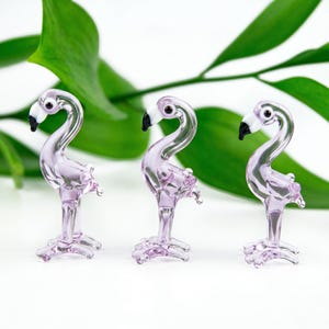 May include: Three small, lavender-coloured glass flamingo figurines. Each has a black beak, white face, and detailed legs and feet. The background features bright green leaves, contrasting with the delicate glass figures. A decorative item.