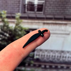 May include: A small, dark green dagger with a curved blade and a cross-shaped hilt. The miniature weapon is held on a fingertip, showcasing its size and detail. The background is blurred, suggesting an outdoor setting.