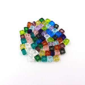 Può includere: Una collezione di perline quadrate colorate in vari colori, tra cui verde, blu, viola e arancione. Le perline sono traslucide e sembrano essere fatte di vetro o materiale simile. Sono sparse su una superficie bianca.