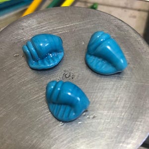 May include: Three turquoise-colored, handcrafted glass seashell beads. Each bead has a detailed, ridged texture, and a glossy finish. The beads are arranged on a silver-colored metal surface, with colorful glass rods visible in the background.