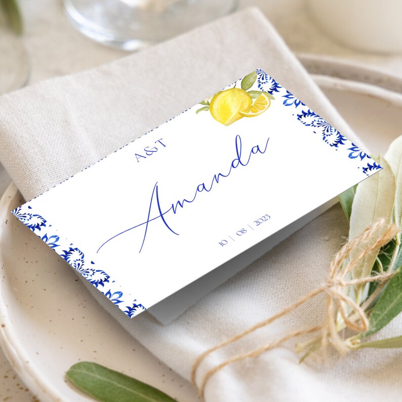 May include: A white place card with blue and white patterned border. The card has the text "A&T" and "Amanda" in blue script. The date "10 | 08 | 2023" is printed in blue at the bottom of the card. A lemon illustration is in the top right corner.