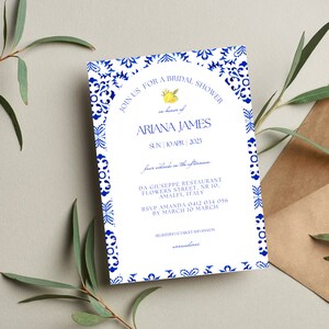 May include: A white bridal shower invitation with a blue and white patterned border. The invitation features a lemon illustration and the text "JOIN US FOR A BRIDAL SHOWER in honor of ARIANA JAMES SUN | 10 APR | 2023 four o'clock in the afternoon DA GIUSEPPE RESTAURANT FLOWERS STREET, NR 10, AMALFI, ITALY RSVP AMANDA 0412 034 056 BY MARCH 10 MARCH REGISTERED AT EMZIGE AND AMVON arrivederci"