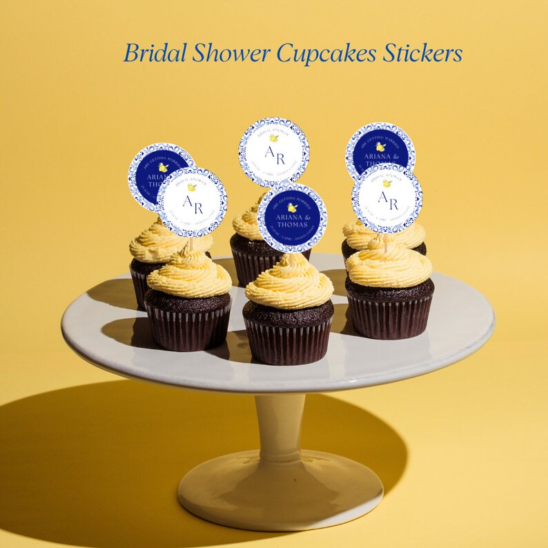 May include: Six cupcakes with yellow frosting and blue and white cupcake toppers with the text "Ariana & Thomas" and "AR" on them. The cupcakes are on a white cake stand.